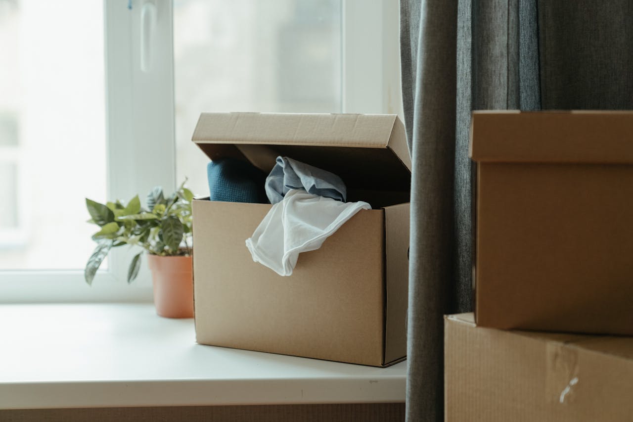 why-choose-us Open cardboard box with clothes by window, symbolizing moving or relocation.