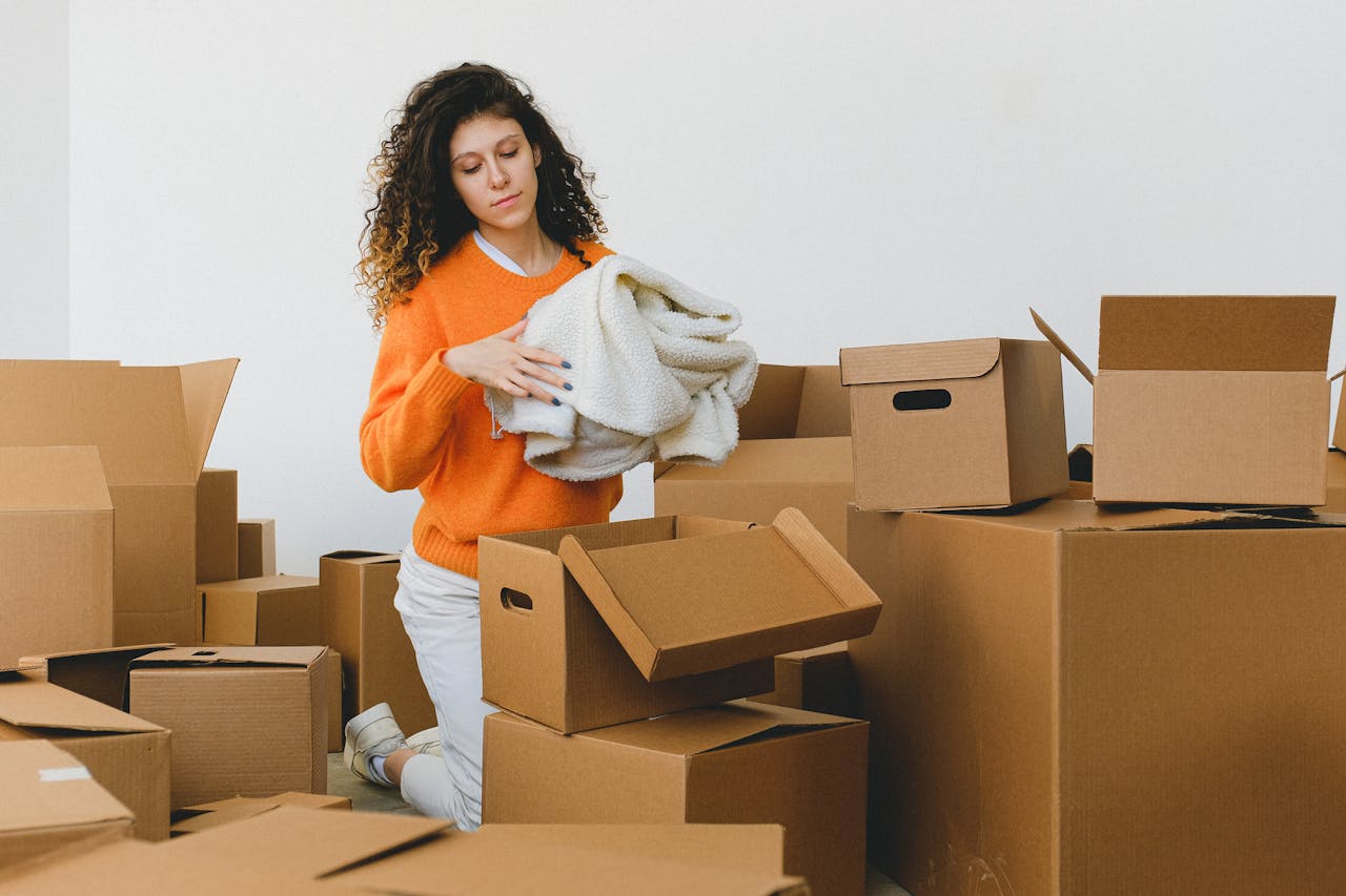 about-02 Concentrated young lady with curly hair standing on knees on floor and packing clothes into carton boxes before moving into new flat